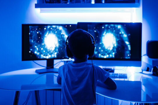 Back view of unrecognizable male kid in headphones watching film about planets on desktop computer while sitting at table in room with blue light