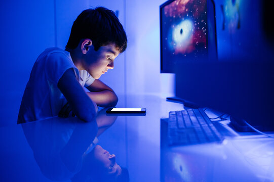 Side view of confused boy watching cellphone empty screen and touching head while sitting in room with blue light at home