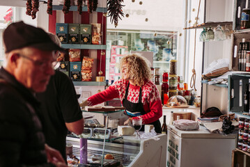 Senior man choosing food from freezer while buying wine and cheese from female seller in cozy small delicatessen store