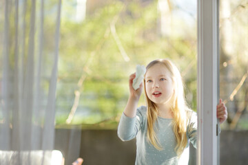 Cute young girl cleaning a window with paper cloth. Child helping with everyday chores.