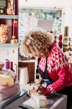 Adult woman in apron slicing fresh cheese while working in cozy local food shop