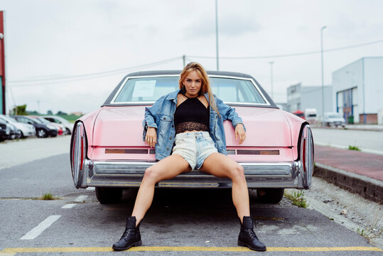 Sensual blonde girl sitting on the bumper of a classic pink car looking at the camera