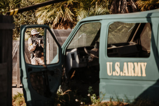 Man in camouflage hiding behind wooden barrier and shooting airsoft gun behind military vehicle while playing tactical game