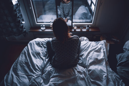 Back view of unrecognizable depressed young female sitting on bed in dark bedroom looking at mirror at home