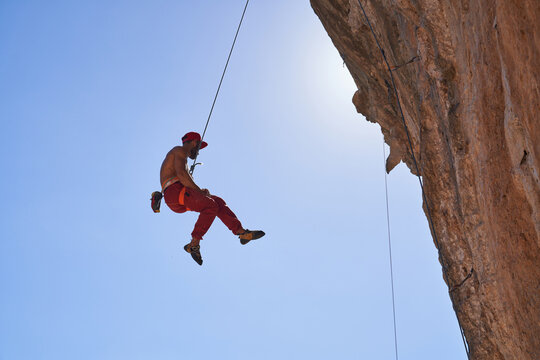From below of strong shirtless bearded mountaineer in red cap hanging on rope while ascending rocky slope of mountain on sunny cloudless day