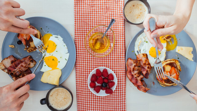 From above view of crop anonymous couple eating tasty fried eggs with cheese and bacon and pancakes with berries for breakfast at home