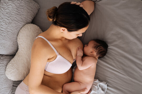 From above of peaceful female in white underwear lying down on comfortable bed and breastfeeding little cute baby