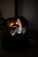 Pensive female in casual t shirt sitting in dark cozy room near fireplace and reading book on netbook while resting during night time