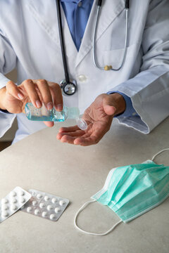 Male therapist in medical gown sitting at table in hospital and sanitizing hands with antiseptic while preparing for treatment of patients during coronavirus outbreak