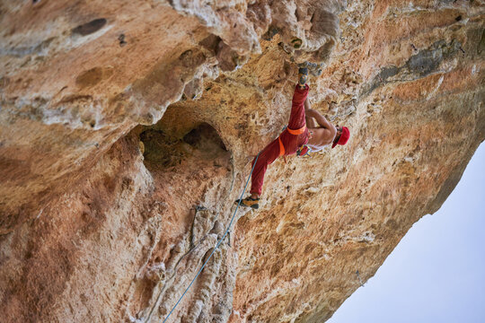 From below of strong shirtless bearded mountaineer in red cap climbing ascending rocky slope of mountain on sunny cloudless day