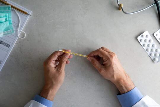 Top view of hands of elderly physician sitting at table in clinic and checking thermometer while working in hospital during coronavirus pandemic