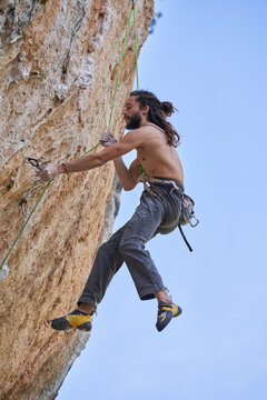 From below side view of shirtless bearded alpinist in safety equipment climbing up on rocky slope of mountain and looking up
