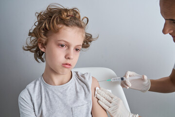 Crop nurse in white latex gloves holding shoulder of boy with curly hair while giving vaccine injection with syringe in modern clinic
