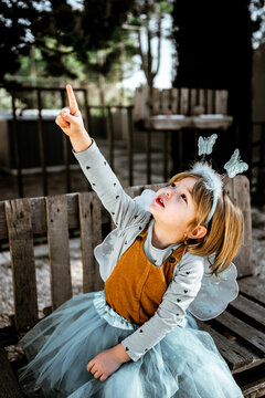 Adorable little girl in fairy costume sitting on shabby wooden bench and looking at camera while spending time in yard