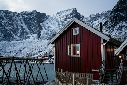 Cottage with red striped walls and white window frames on wooden pier on strait shore against snowy township at foothill in winter cloudy day in Lofoten