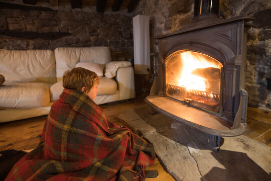 Cuddling kid wrapped in cozy tartan plaid sitting on wooden floor near burning fireplace and looking at fire in stone house in Cantabria