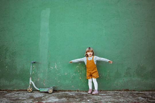 little girl with outstretched arms standing near weathered green wall and kick scooter and counting while playing hide and seek
