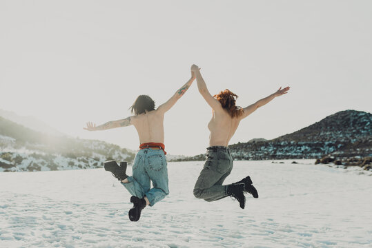 Back view of cheerful topless young women in jeans holding hands and jumping in snowy field in sunny winter day with mountains in background