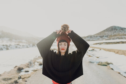 From below of happy active young female dressed in hat with fur pompom and sweater with jeans having fun and enjoying nice sunny winter day at countryside