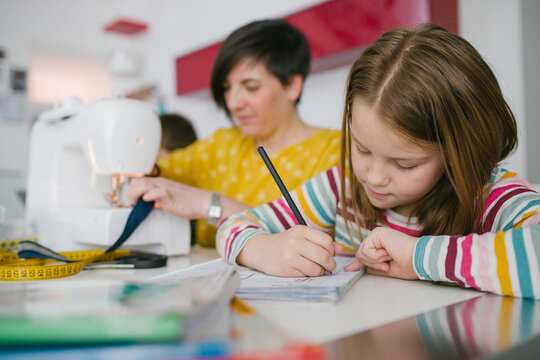 Focused girl doing homework assignment while sitting near adult woman sewing garment at home