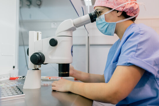Side view of woman in medical uniform and mask using modern microscope to examine human cells while working in lab of modern clinic