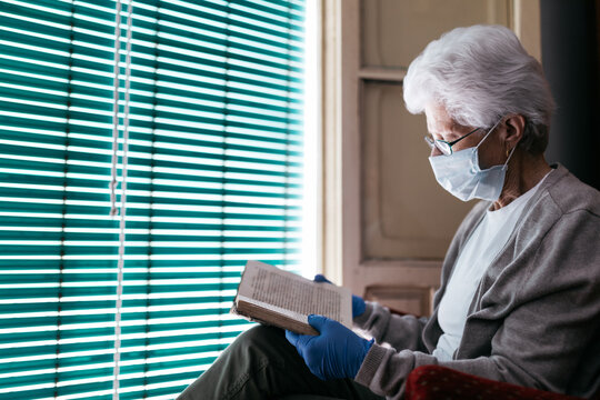 Side view of lonely elderly woman in medical mask and protective gloves sitting in front of shuttered window and reading book while spending day at home during self isolation because of coronavirus