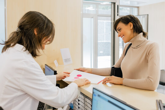 Smiling specialist in white uniform and with name tag reviewing documents while conversing with unrecognizable female patient at reception desk in clinic