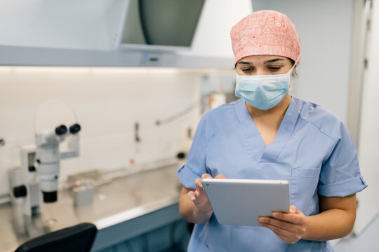 Woman in medical uniform and mask browsing data on tablet while working in contemporary lab in clinic