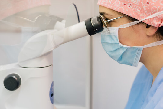 Side view of woman in medical uniform and mask using modern microscope to examine human cells while working in lab of modern clinic