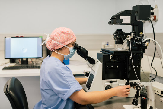 Side view of woman in medical uniform and mask using modern machine to fertilize egg cell during work in laboratory of modern fertility clinic