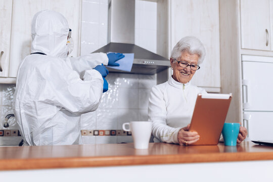 Unrecognizable home care worker in protective uniform cleaning kitchen while cheerful elderly woman enjoying new book during self isolation at home due to coronavirus pandemic