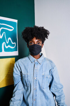 Young African American female in casual denim shirt and black protective mask looking at camera while staying at home during coronavirus outbreak