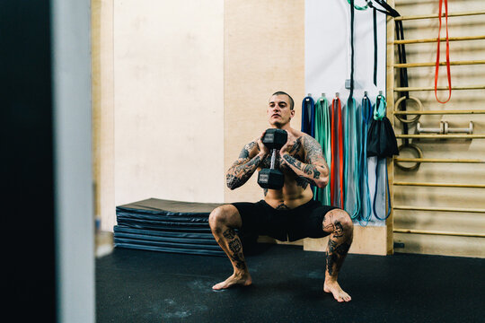 Brutal sportsman with naked torso squatting with dumbbell during workout in contemporary gym
