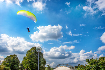 Gliding using a parachute over green trees in blue cloudy sky over a park.