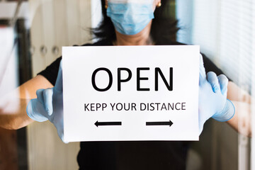 woman puts an "open, keep your distance" sign on the door of a business. with reflections of the glass.