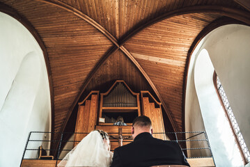 Rear view of marriage couple in front of altar in church. Focused on woman playing at pipe organs....