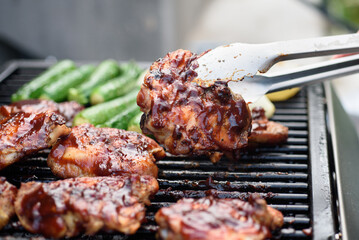 Flipping a barbecued chicken thigh with tongs on a gas grill with baby zucchini in the background.