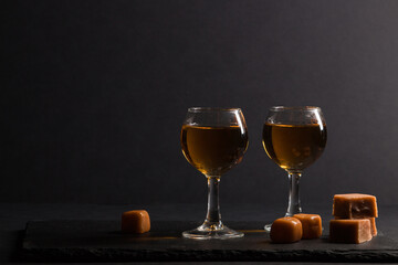Glass of whiskey with caramel candies on a black stone slate board on black background. Side view, low key, copy space.