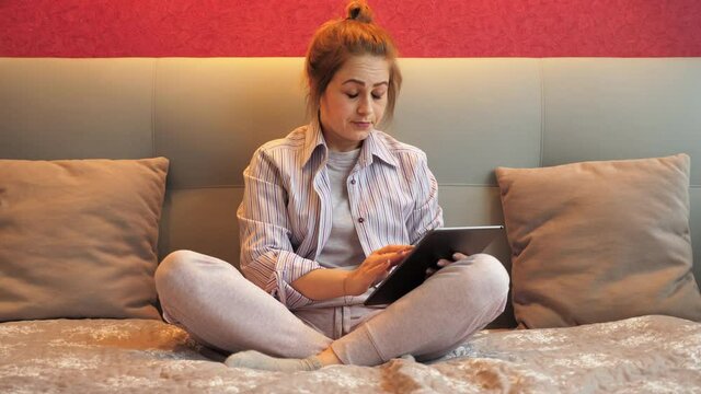 Tired brunette woman at home sitting on the bed uses the tablet for work, yawns and falls asleep. 4K footage