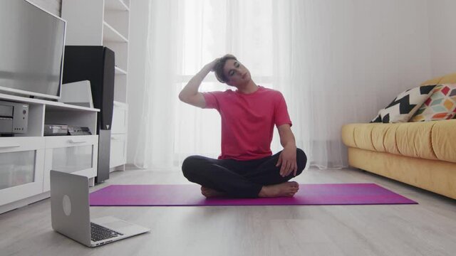 Young transgender homosexual man with make up stratching neck on mat at home with help of his instructor online on laptop at home