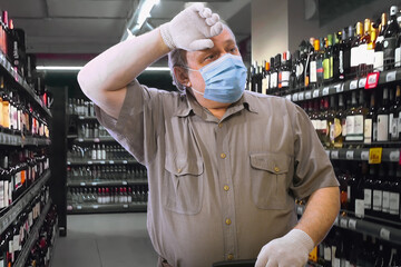 An elderly man in a medical mask and white gloves, wiping the sweat from his forehead with his hand, searches for medicine in a host of bottles of alcohol on the shelves of the store