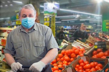 An elderly man in a short-sleeve shirt, medical mask and white gloves stands near boxes with red tomatoes in the market.