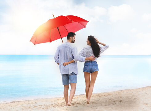 Happy young couple with umbrella for sun protection walking on beach near sea