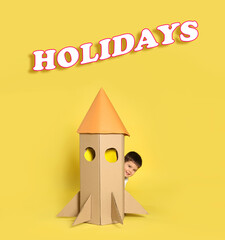 School holidays. Cute little child playing with cardboard rocket near yellow wall