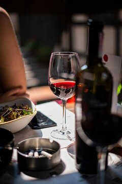 Vertical image of exquisite glass of red wine standing on a table next to bottle, ashtray with cigarettes and plate of salad in restaurant, bar, cafe on the terrace on a sunny summer day with a woman