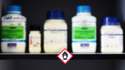 Chemical Hazard Sign pictogram, Globally Harmonized System of Classification and Labelling of Chemicals (GHS) Flammable substance category with container bottles on shelf cabinet, blurred background.