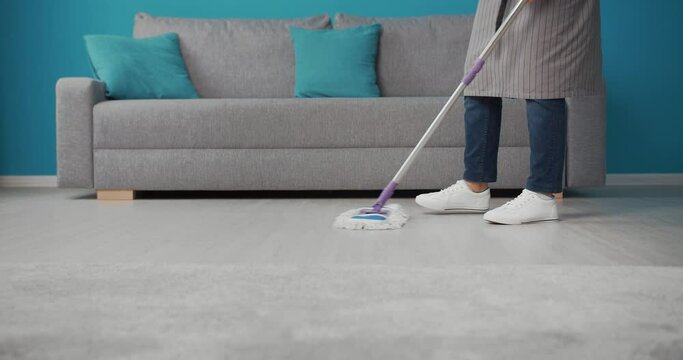 Housewife in grey apron and white sneakers using swabber for washing floor at living room. Mature lady doing regular hygiene for maintenance cleanliness at home.