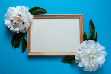 Flower composition. Wooden photo frame and beautiful white peonies (Paeonia) with green leaves on a pastel blue background.