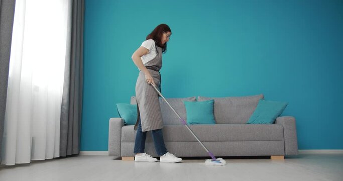 Side view of mature woman in domestic outfit and grey apron cleaning floor with mop. Concept of people, housework and housekeeping.