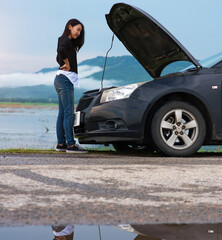 Asian woman with a broken car with open hood.Young woman using mobile phone while looking at broken down car on street